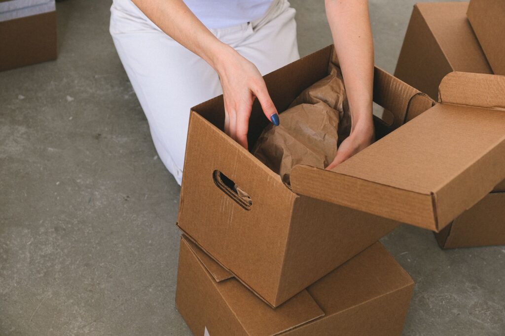 Hands placing a brown-paper-wrapped item inside an open cardboard box.