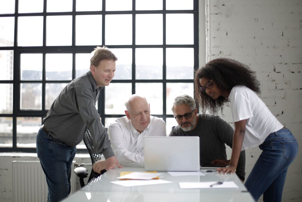 Four colleagues gathered around a laptop on a table beside large windows.