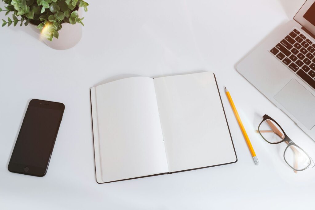 Open blank notebook beside a laptop, phone, pencil and glasses on a white desk.