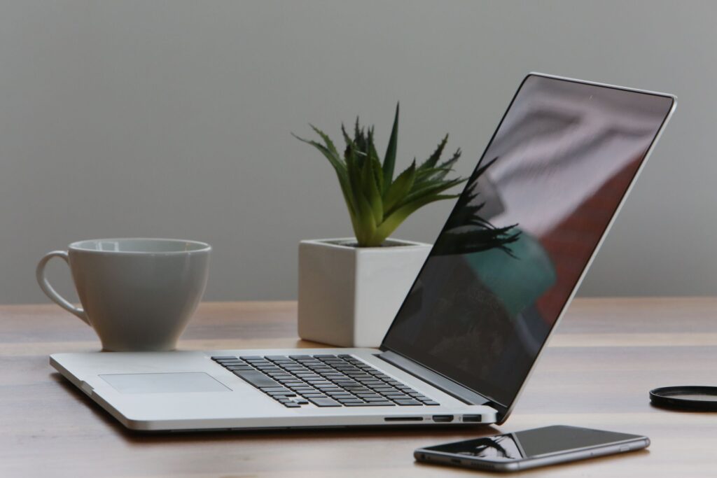 Open laptop beside a cup, potted plant and phone on a wooden desk.