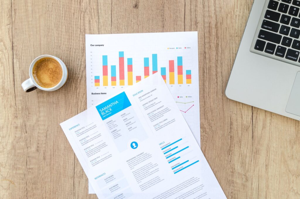 Printed charts and a CV beside a laptop and cup of coffee on a wooden desk.
