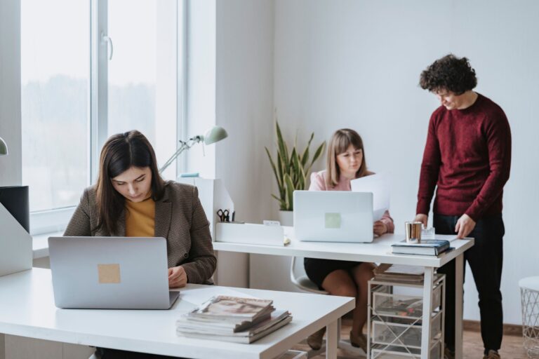 Three people working with laptops and paperwork at office desks.